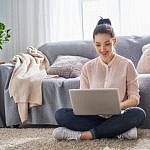 Woman sitting cross-legged on a carpeted floor, typing on a laptop. Shes in a cozy living room with a gray sofa, cushions, and a beige throw in the background. Shes smiling and casually dressed, with her hair in a bun. Natural light fills the room.
