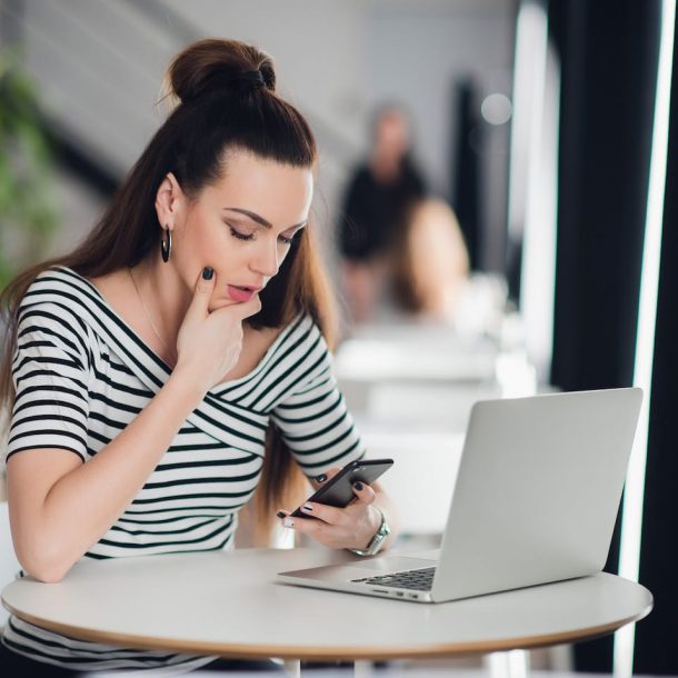 A woman with long hair is sitting at a round white table in a cafe, looking at her phone. She is wearing a black and white striped top. A laptop is open in front of her. The background is softly blurred.