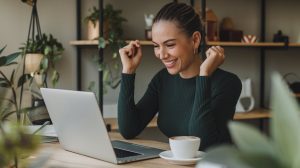 A woman sits at a wooden table, beaming with excitement as she looks at her laptop, fists raised in delight. With a fresh cup of coffee beside her and shelves adorned with plants and decorations nearby, shes ready to embark on the exciting journey of starting a blog.
