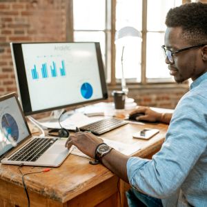 A person sits at a wooden desk, diligently working on a laptop and desktop computer displaying graphs and charts for Bloom Digital. They wear glasses and a blue shirt. The background shows large windows with natural light streaming in.