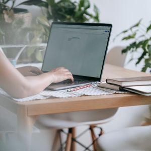 A person types on a laptop at a wooden table surrounded by green plants. The table holds a few books and pens, with the ambiance of Bloom Digital seamlessly enhancing the experience. Bright, natural light illuminates the scene, creating a calm and focused atmosphere.