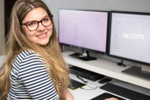 Woman with long blonde hair and glasses smiling at the camera, sitting at a desk with two monitors displaying digital content. She wears a striped shirt and is in a bright, modern workspace.