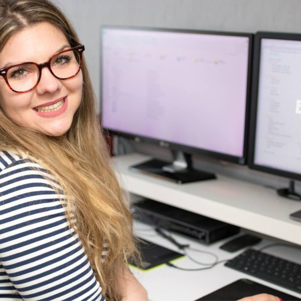 Woman with long blonde hair and glasses smiling at the camera, sitting at a desk with two monitors displaying digital content. She wears a striped shirt and is in a bright, modern workspace.