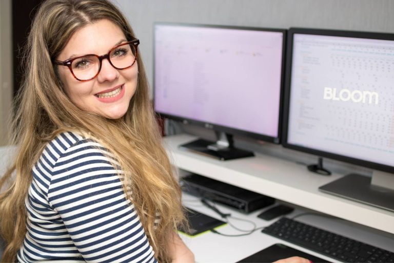 Woman with long blonde hair and glasses smiling at the camera, sitting at a desk with two monitors displaying digital content. She wears a striped shirt and is in a bright, modern workspace.