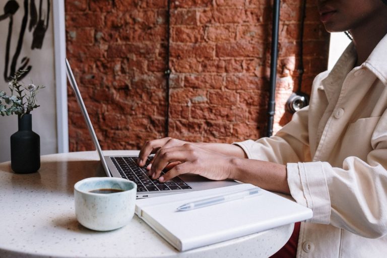 A person in a light-colored jacket types on a laptop at a white round table, perhaps pondering questions about blogging. Next to the laptop, theres an open notebook with a pen and a light green cup. A small black vase with flowers sits in the background against a brick wall.