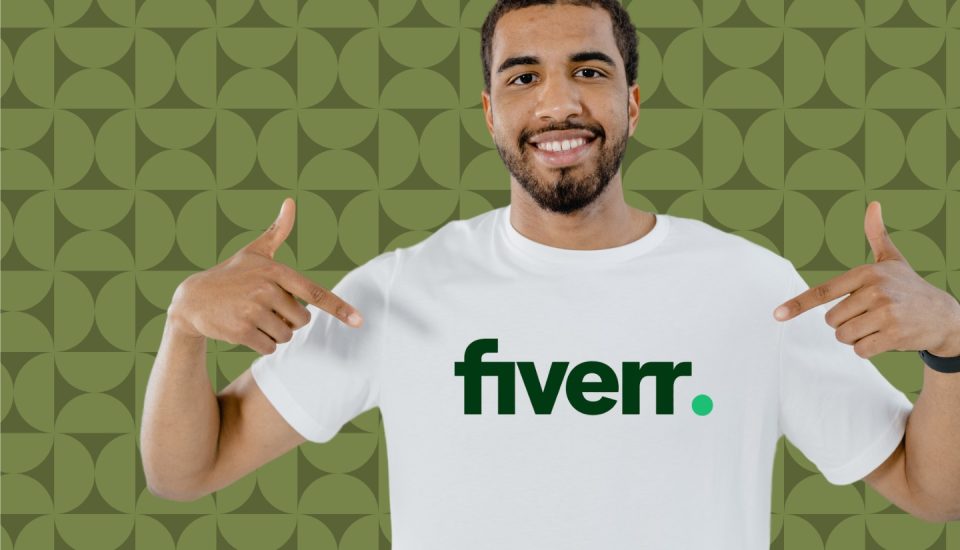 A smiling man points to the Fiverr logo on his white T-shirt, hinting at ways to make money on Fiverr. The background complements him with a green geometric pattern.