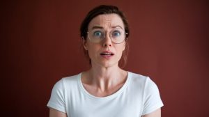 A person with glasses and short brown hair looks surprised against a dark brown background. They are wearing a white shirt.