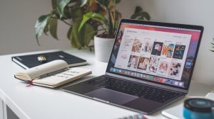 A laptop displaying a website is on a desk, perfect for researching what to pin on Pinterest. An open notebook, a closed notebook, and a pen lie nearby. In the background, theres a potted plant and wall plug, completing this cozy home office setup.