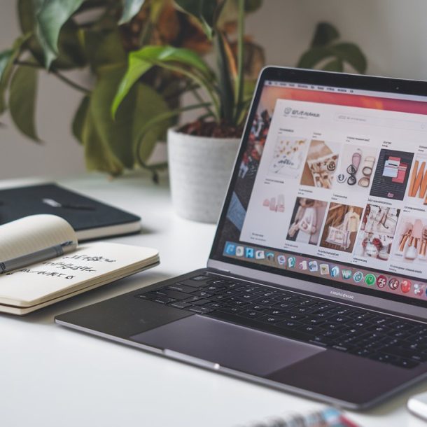 A laptop displaying a website is on a desk, perfect for researching what to pin on Pinterest. An open notebook, a closed notebook, and a pen lie nearby. In the background, theres a potted plant and wall plug, completing this cozy home office setup.