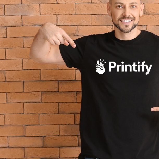 A man smiling and pointing at his black t-shirt with the Printify logo. He stands in front of a brick wall background.