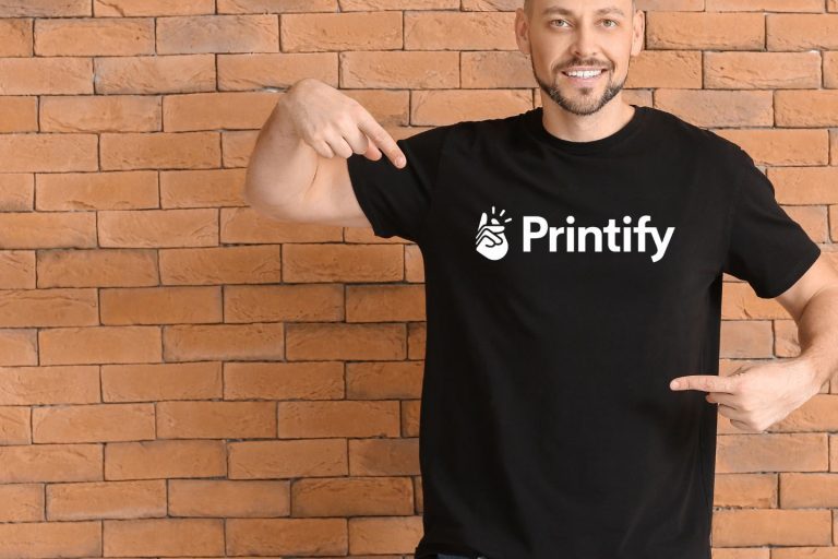 A man smiling and pointing at his black t-shirt with the Printify logo. He stands in front of a brick wall background.