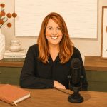 A woman with red hair smiles while sitting at a desk with a microphone. The desk has books, plants, a notebook, and décor. Art and a vase with orange flowers are displayed in the background.