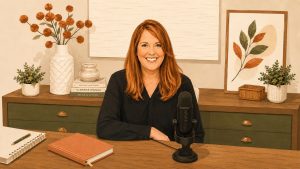 A woman with red hair smiles while sitting at a desk with a microphone. The desk has books, plants, a notebook, and décor. Art and a vase with orange flowers are displayed in the background.